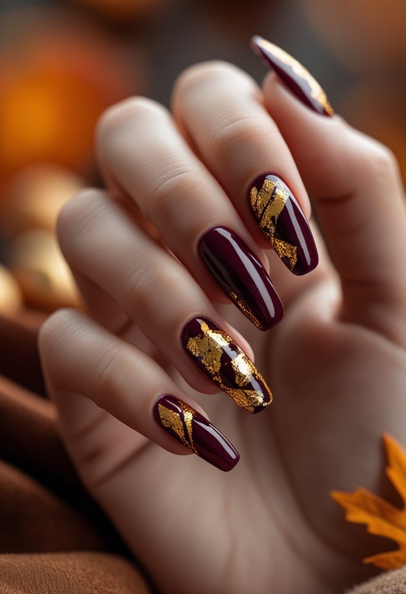 Close-up of a hand with deep burgundy nails accented with gold foil, against a warm, blurred autumn background.